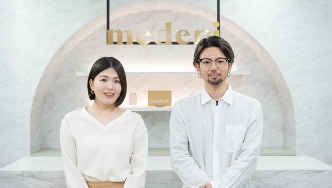 左から阿部様、星野様がmederiロゴの前に並んで立ち、こちらにむかってほほ笑んでいる写真
