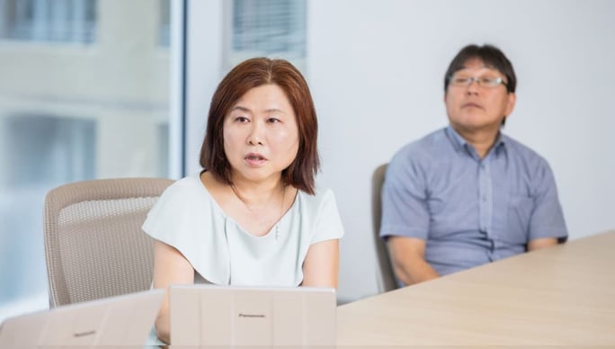 オフィスの会議室でノートパソコンを前にした女性が真剣な表情で話している。背後には別の男性が座り、話を聞いている。