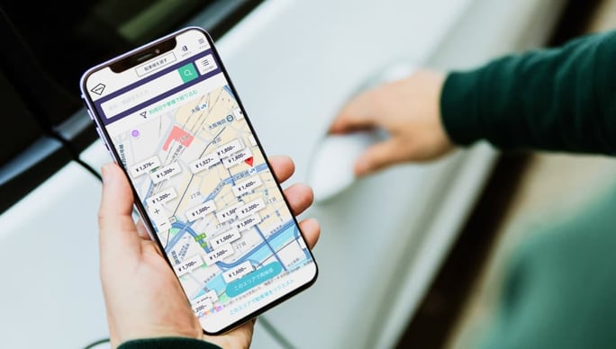 スマートフォンのakippaアプリで駐車場を検索し、料金や場所を確認している様子。車のドアを開けようとしている様子（画面）