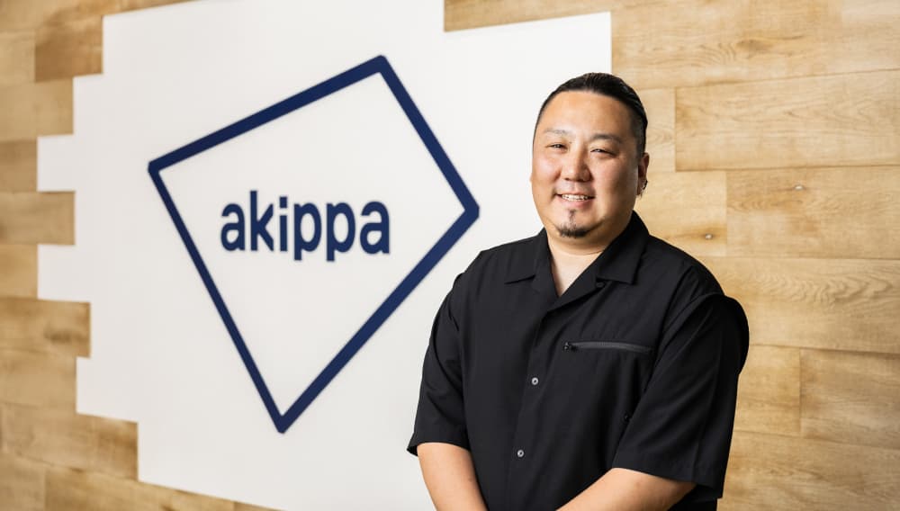 akippaロゴの前で微笑む荒木様(写真)