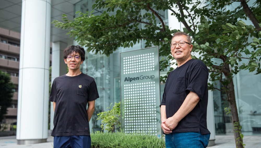 アルペングループのロゴ看板前に立つお二人の写真