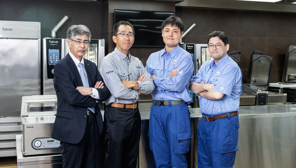 業務用厨房機器が並ぶフジマックのキッチンで、スーツ姿の男性1名と作業着姿の男性3名が腕を組んで並んでいる集合写真