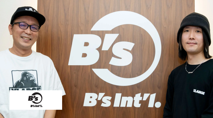 「B's B's Int'l. 」と書かれた壁の左右にキャップを被った白いTシャツの男性とバケットハットを被った黒いTシャツの男性が立っている写真。左下に「B's」のロゴがある。