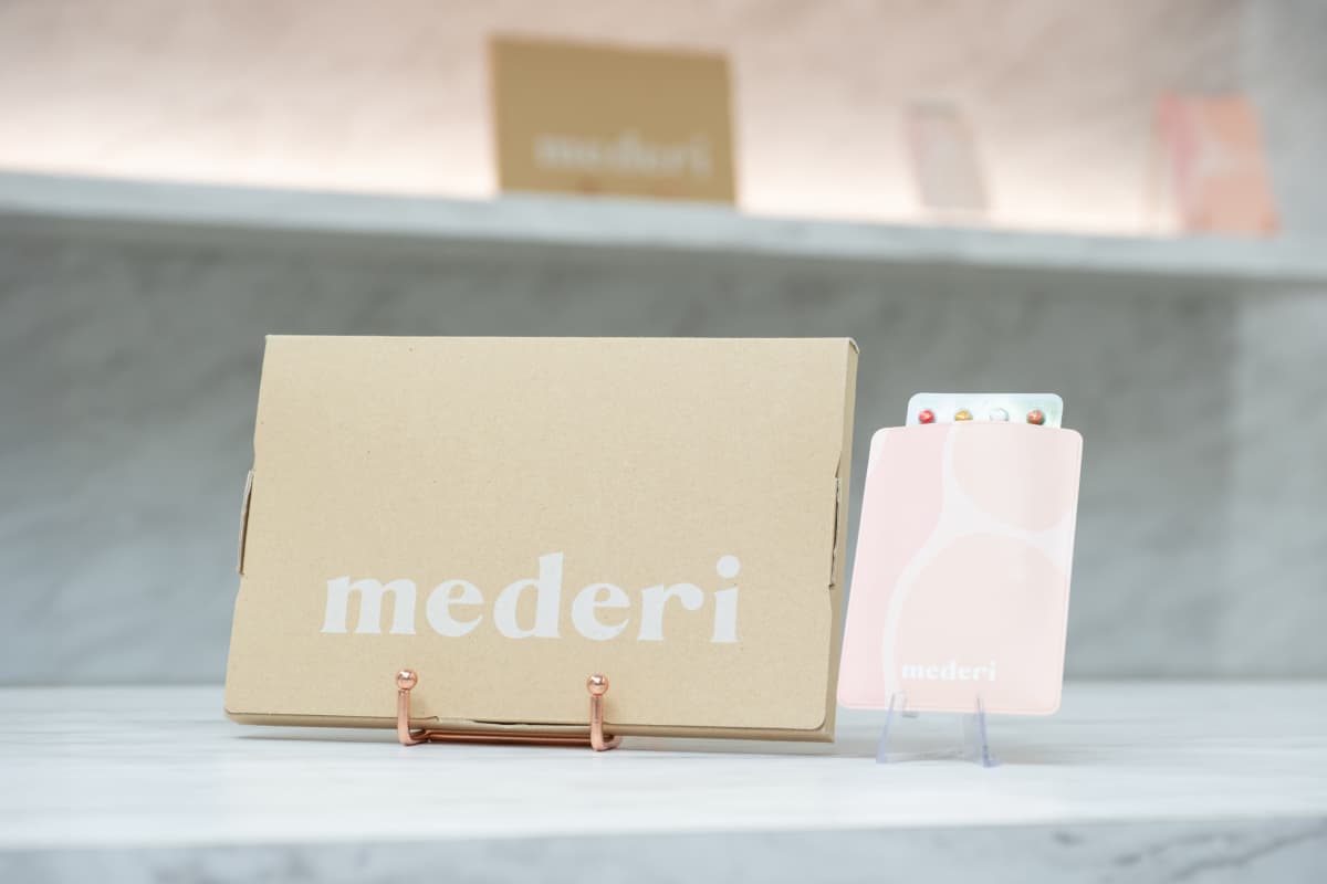 左にmederiのロゴ入りのクラフト紙パッケージ、右にピンク色のケースに入ったカラフルなピルシートが並んで配置されている写真