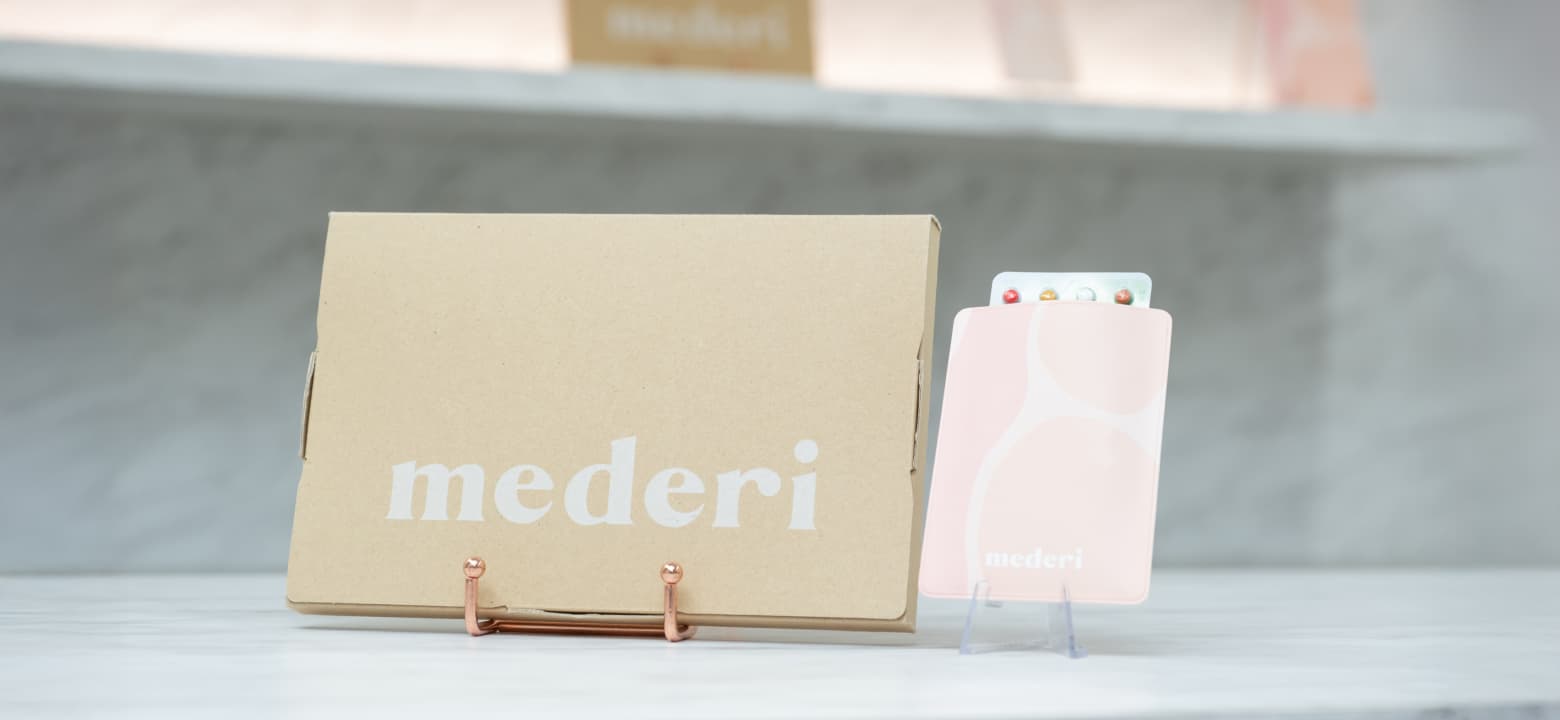 左にmederiのロゴ入りのクラフト紙パッケージ、右にピンク色のケースに入ったカラフルなピルシートが並んで配置されている写真