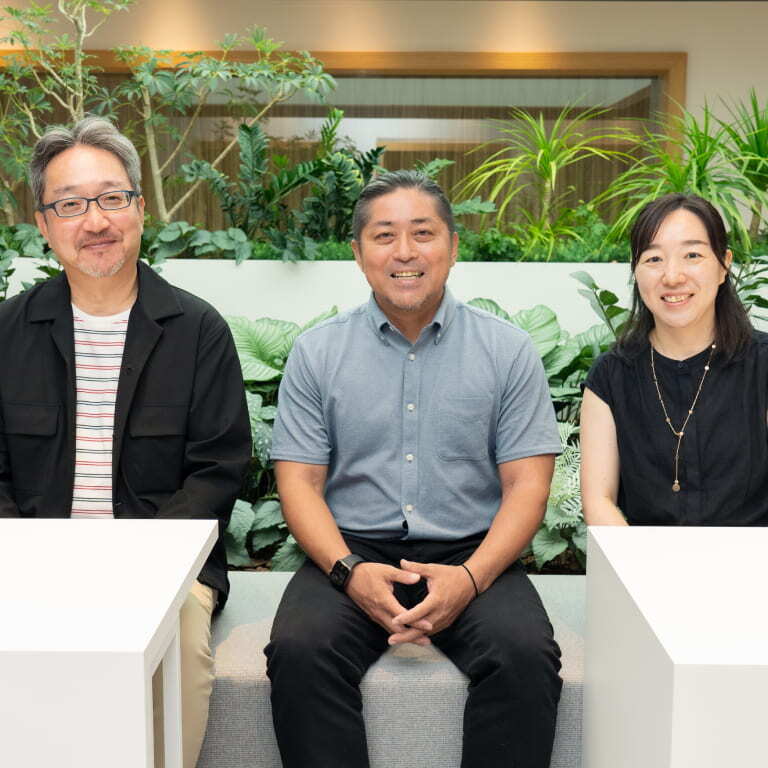 室内のグリーンを背景に、笑顔で座る3人の社員の集合写真