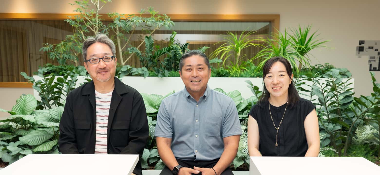 室内のグリーンを背景に、笑顔で座る3人の社員の集合写真