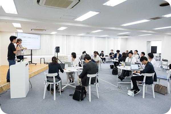 前方の大型スクリーンにスライドが投影され、多くの参加者が円卓を囲んでいる会場全体の様子