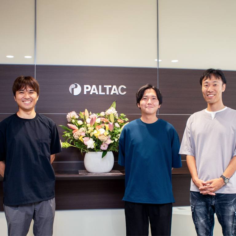 PALTACと書かれた壁の前に3名の男性が並んでいる